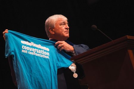 Dr. Schrader holds blue t-shirt with white text that reads "Conversation not Confrontation."