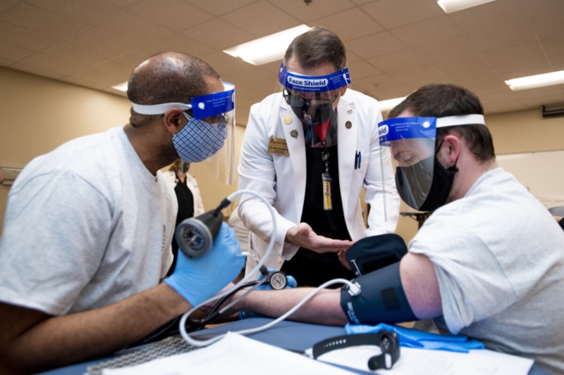 Physician assistants in masks and face shields practice taking blood pressure.